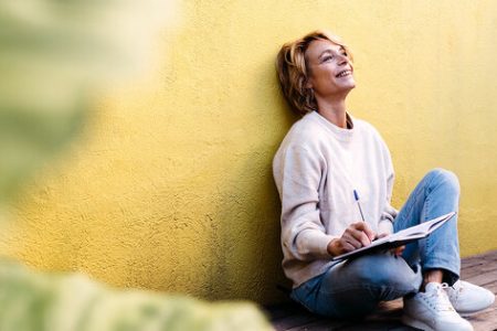 Frau sitzt an einer Hauswand, schreibt etwas in ein Buch und schaut glücklich nach oben. Joyce Meyer